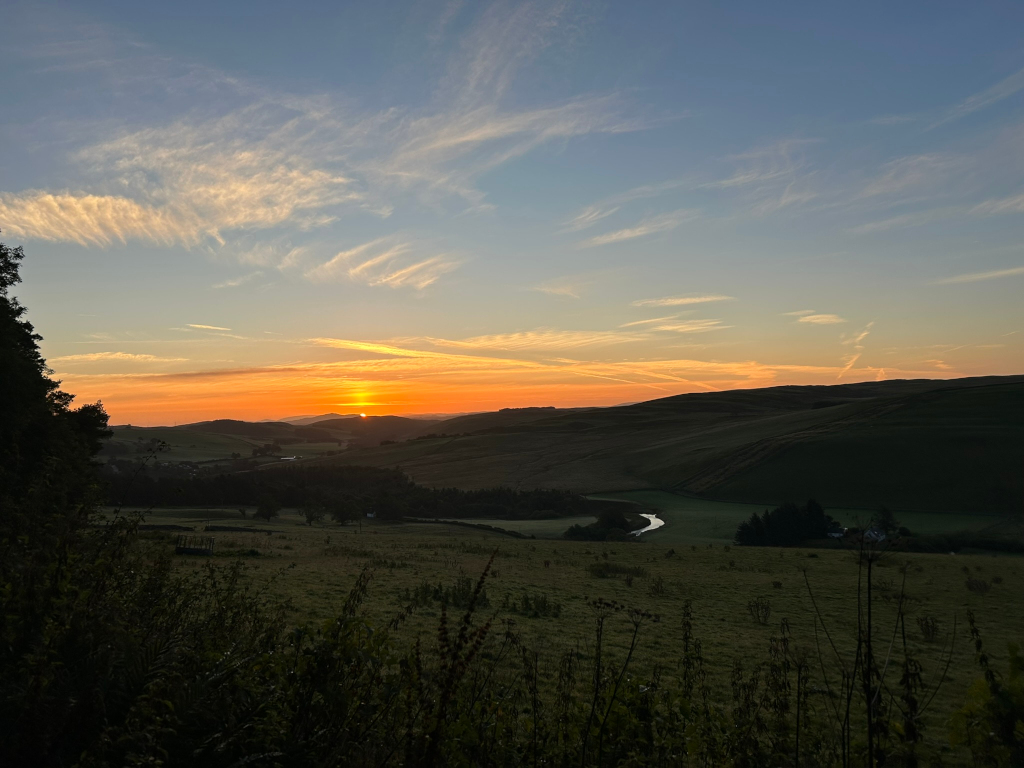 Breathtaking sunrise over a rolling landscape. The sun, a vibrant orange, is partially visible on the horizon, casting a warm glow across the undulating hills and valleys. The sky is a blend of soft oranges, pinks, and blues, with wispy clouds adding texture and depth. A calm river meanders through the valley below, providing a peaceful counterpoint to the dramatic sky. The foreground consists of dark, silhouetted vegetation, contrasting with the brightly lit landscape in the distance.