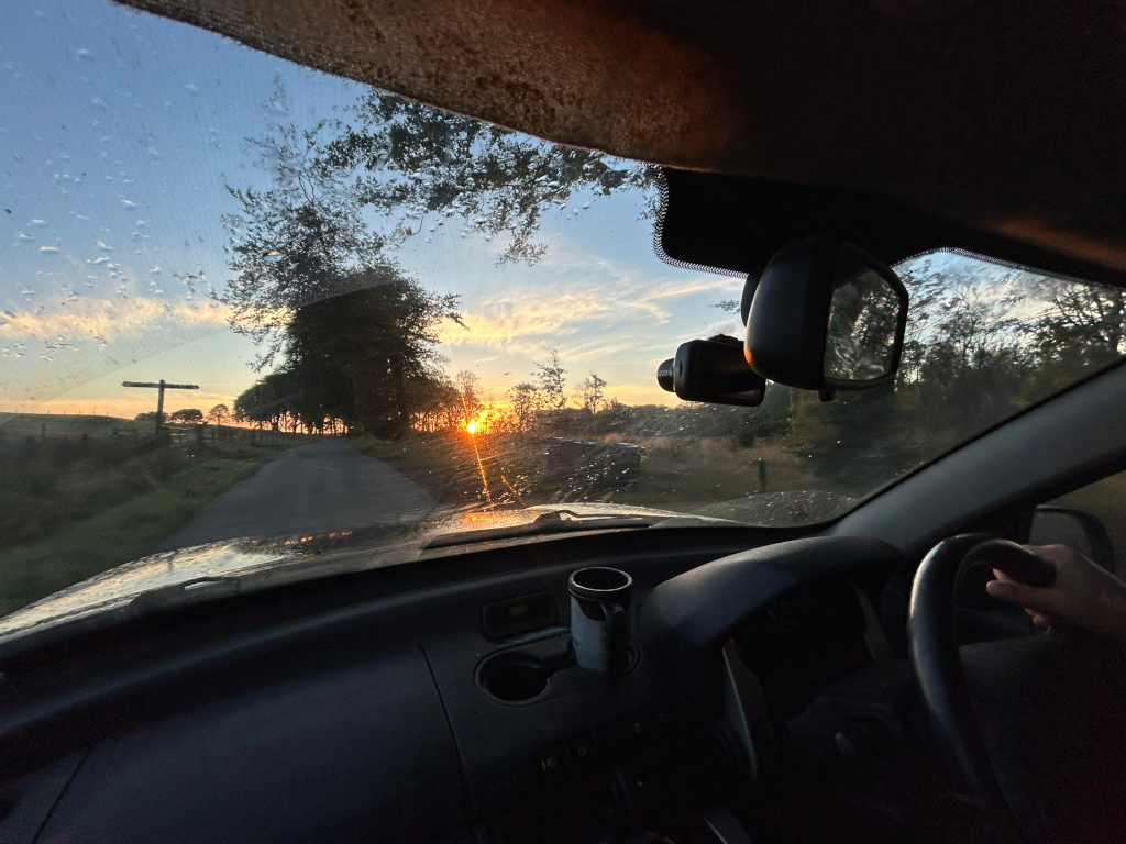 First-person view from inside a car driving down a country road at sunrise. The sun is rising on the right side of the frame, casting a warm glow on the landscape. The road is lined with trees, and a simple wooden signpost can be seen in the distance. The wind shield is wet with raindrops, adding to the atmospheric feel of the scene. A dash cam and rear-view mirror are visible within the car. The overall mood is peaceful and evocative of a journey.