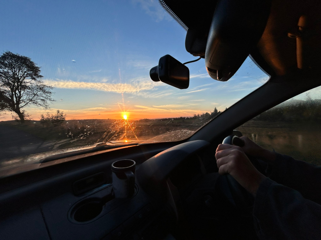 Charlie driving a car at sunrise. The sun is rising in the distance, casting a warm glow over the landscape. The driver's hands are visible on the steering wheel. A cup is in the car's cup holder. The overall mood is peaceful and serene.