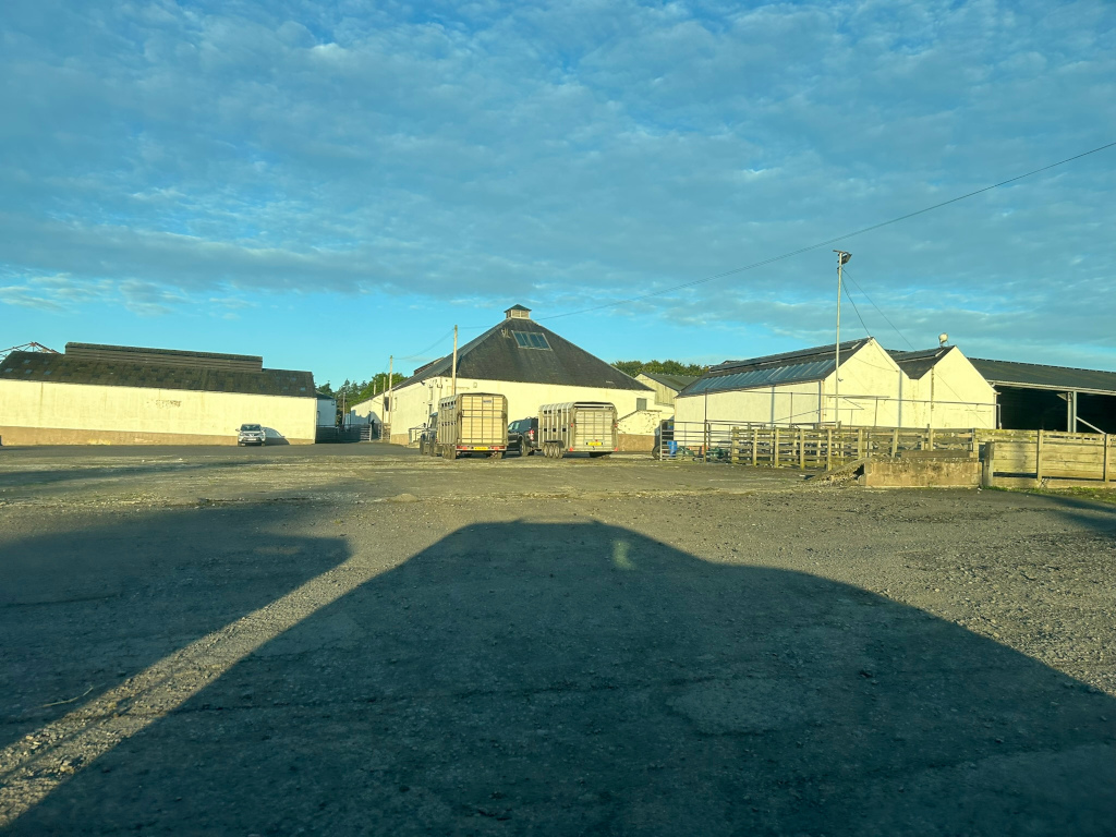 Rural scene, likely a farm or agricultural facility. There are several white buildings, including one with a distinctive conical roof (possibly a grain silo or similar structure). In the foreground is a large, gravelled or paved area. Two livestock trailers are parked near the main building, and a small car is visible in the distance. The overall impression is one of quiet rural industry, under a partly cloudy sky.