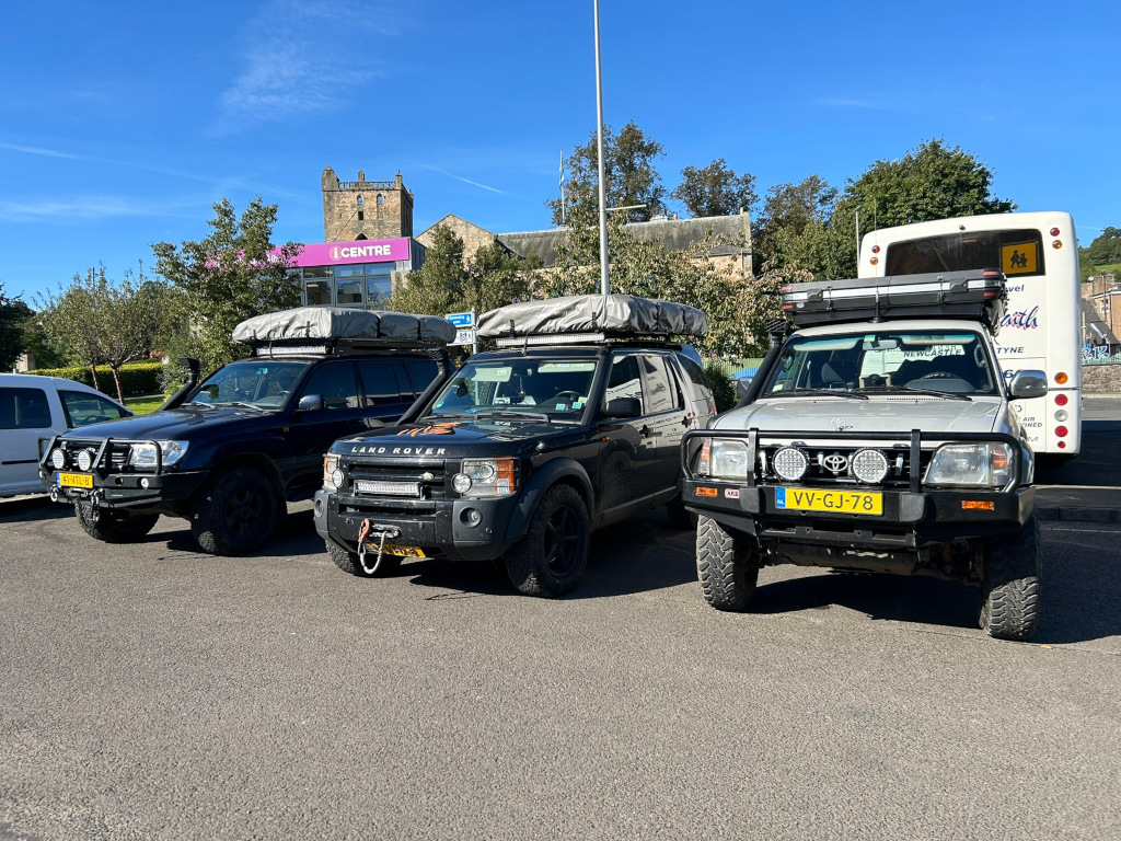Three black and one silver SUVs, all equipped with roof top tents, are parked in a parking lot. In the background is a building that appears to be a church or other historical structure, and a bus. The scene is bright and sunny, suggesting daytime. The vehicles are likely prepared for an overland journey.