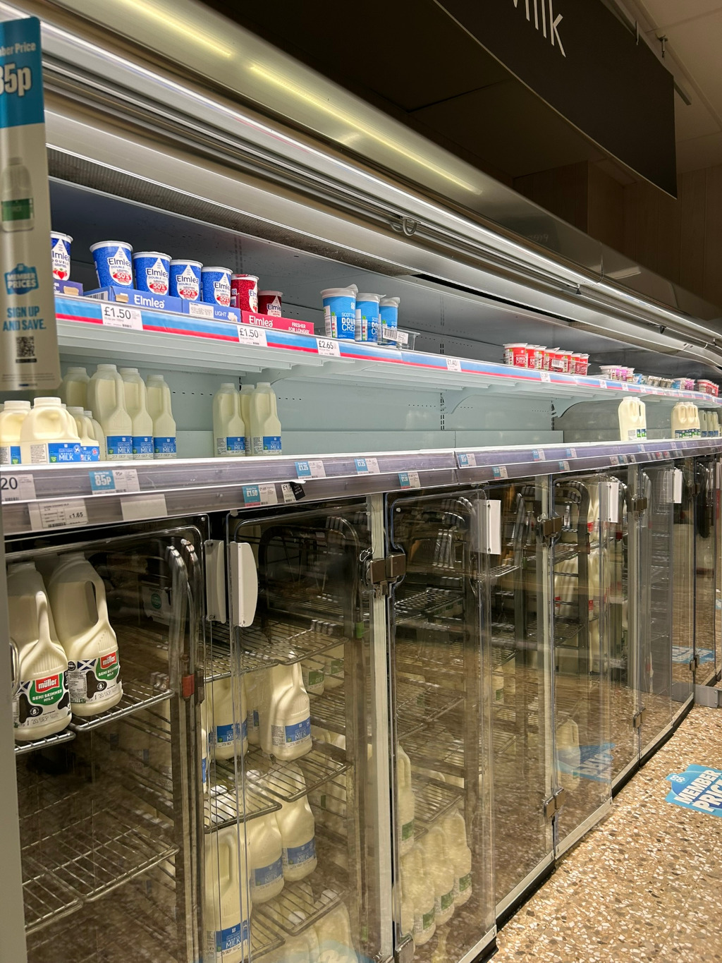 Refrigerated section of a supermarket, specifically a dairy aisle. Multiple glass-fronted refrigerators are filled with various dairy products, primarily milk in various sizes and containers. There are also smaller quantities of yogurts and cream on shelves above the refrigerators. The shelves appear somewhat sparsely stocked, particularly the upper shelves. The image suggests a snapshot of a typical supermarket dairy section, with a focus on the refrigerated milk display. There's also price labelling visible on some items.