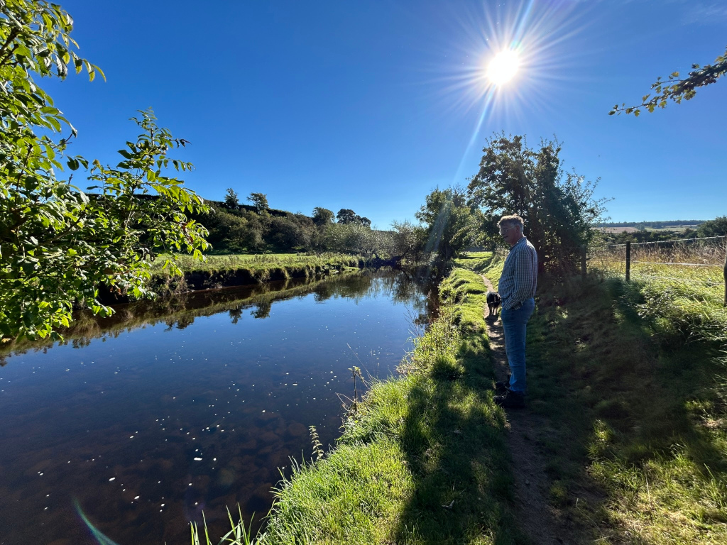 Charlie standing by a calm river on a sunny day. He is accompanied by a small dark-coloured dog. Lush green vegetation lines the riverbanks, with trees visible in the background. The sky is a clear, vibrant blue, and the sun is shining brightly. The overall mood is peaceful and serene, suggestive of a tranquil countryside setting.