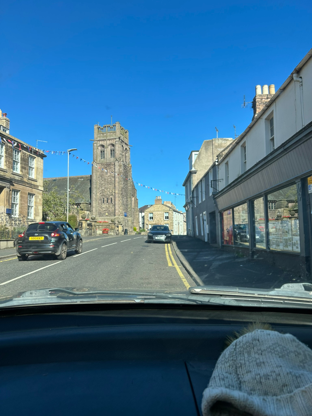 Street scene in a small town or village, likely in the UK, judging from the car license plate. The perspective is from inside a car driving down a street. A stone church tower is prominently featured in the background, with buildings lining the street on either side. Festive bunting is strung across the road, suggesting a celebration or special occasion. The overall impression is one of a quiet, possibly historic, community on a sunny day.