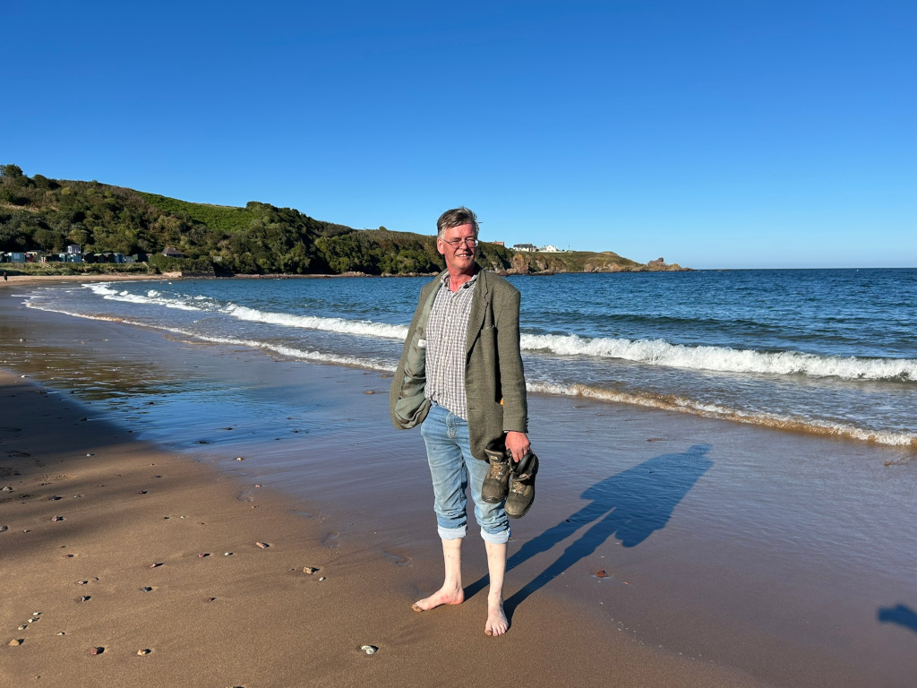 Charlie standing barefoot on a sandy beach, holding a pair of walking boots in his right hand. He is wearing a green jacket and jeans rolled up to his ankles. The beach is calm, with gentle waves lapping the shore. In the background is a green hillside and a clear blue sky.