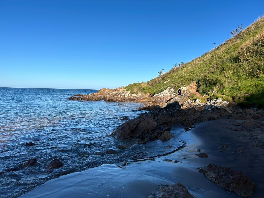 Tranquil coastal scene. The foreground features a dark sand beach merging with the gently lapping waves of a calm sea. Dark rocks are scattered along the shoreline and extend into the water. A verdant, grassy hillside rises gently from the rocks in the background, all under a clear blue sky. 