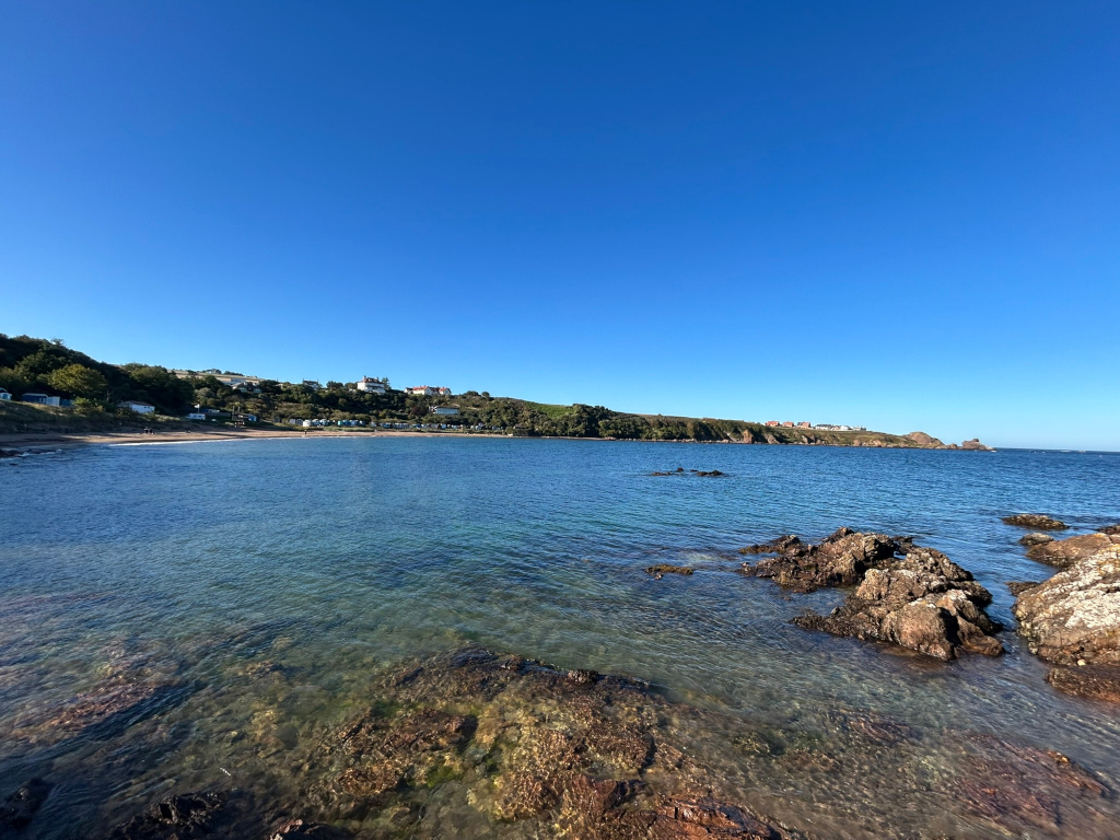 Tranquil coastal scene on a clear, sunny day. In the foreground are clear, shallow waters revealing rocks and some aquatic vegetation. The mid-ground features a calm bay with a sandy beach, sparsely populated. In the background, a gently sloping hillside with low-lying buildings or houses is visible against a deep blue sky.