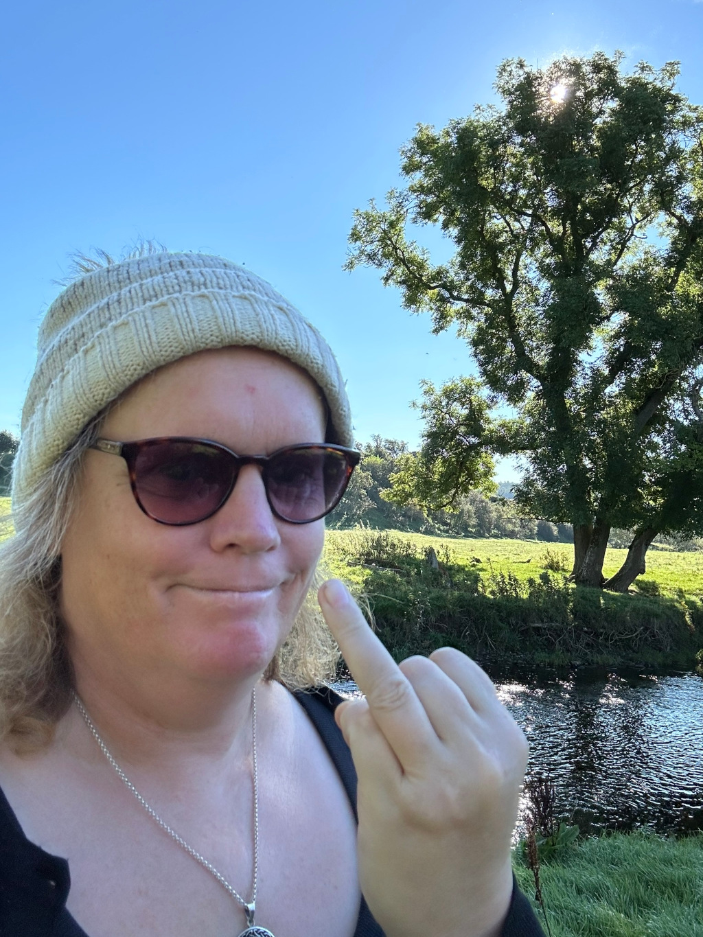 Close-up selfie of Leonie with long blonde hair wearing a beige knit hat and dark sunglasses. She is giving the middle finger to the camera. The background features a tranquil landscape: a calm river, green grass, and a large tree under a bright, sunny sky. The woman's expression is a mixture of amusement and defiance.