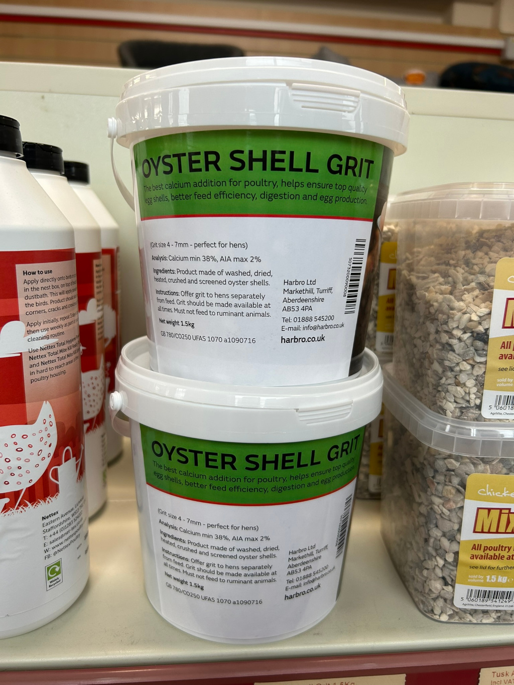 Two buckets of Harbro Oyster Shell Grit for poultry. The buckets are identical, showing product information including grit size, analysis, ingredients, instructions for use, net weight, and contact information for the manufacturer. A partially visible third container of a similar product and a chicken feed mix is also in the image. The overall setting appears to be a retail environment, likely a feed store or agricultural supply shop.