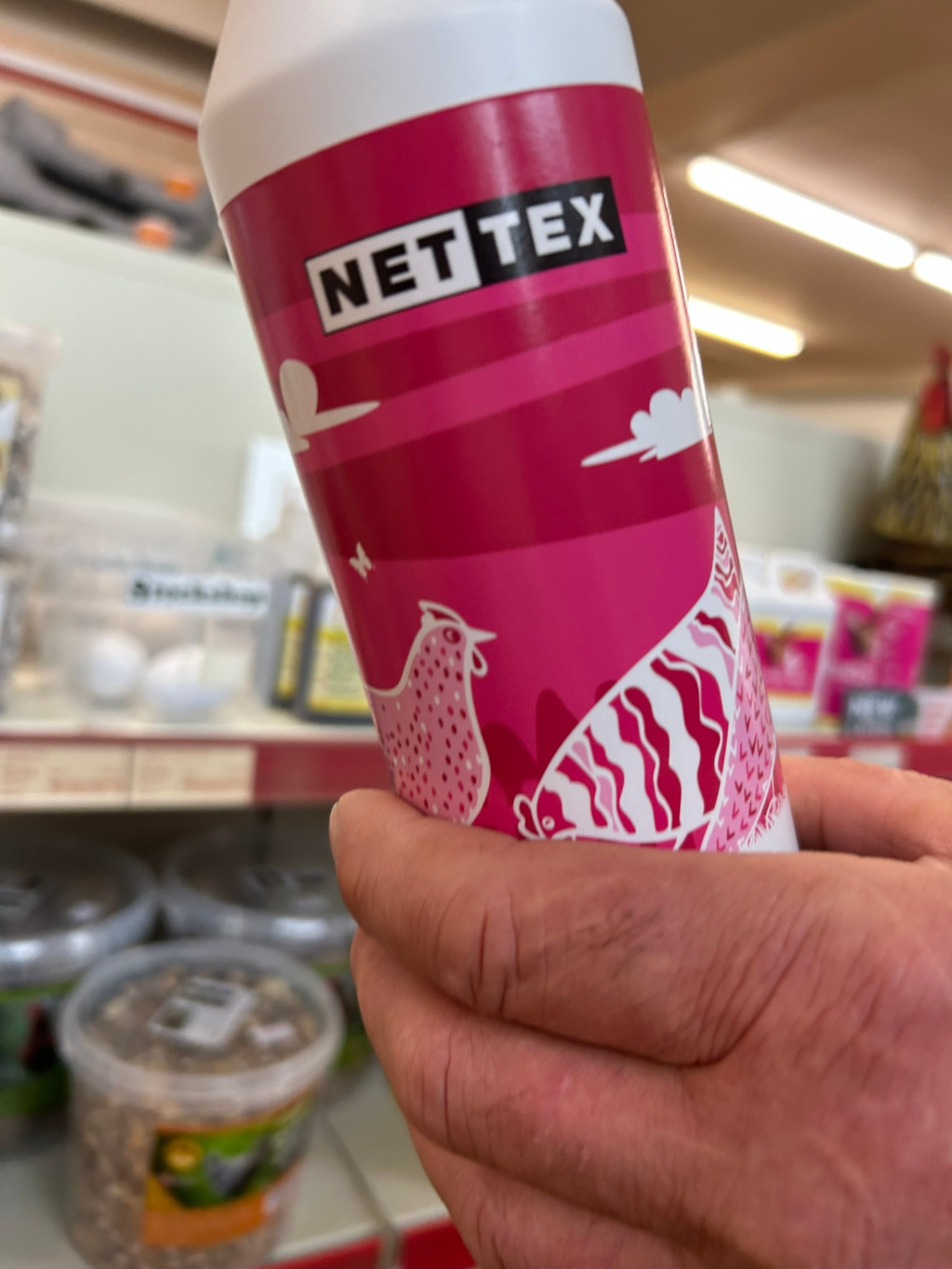 Leonie's hand holding a pink bottle of NETEX product. The bottle features a stylised illustration of chickens and birds against a pink and white background. The bottle is being held in a store aisle with various pet supplies visible in the background, including containers of what looks like birdseed and eggs.
