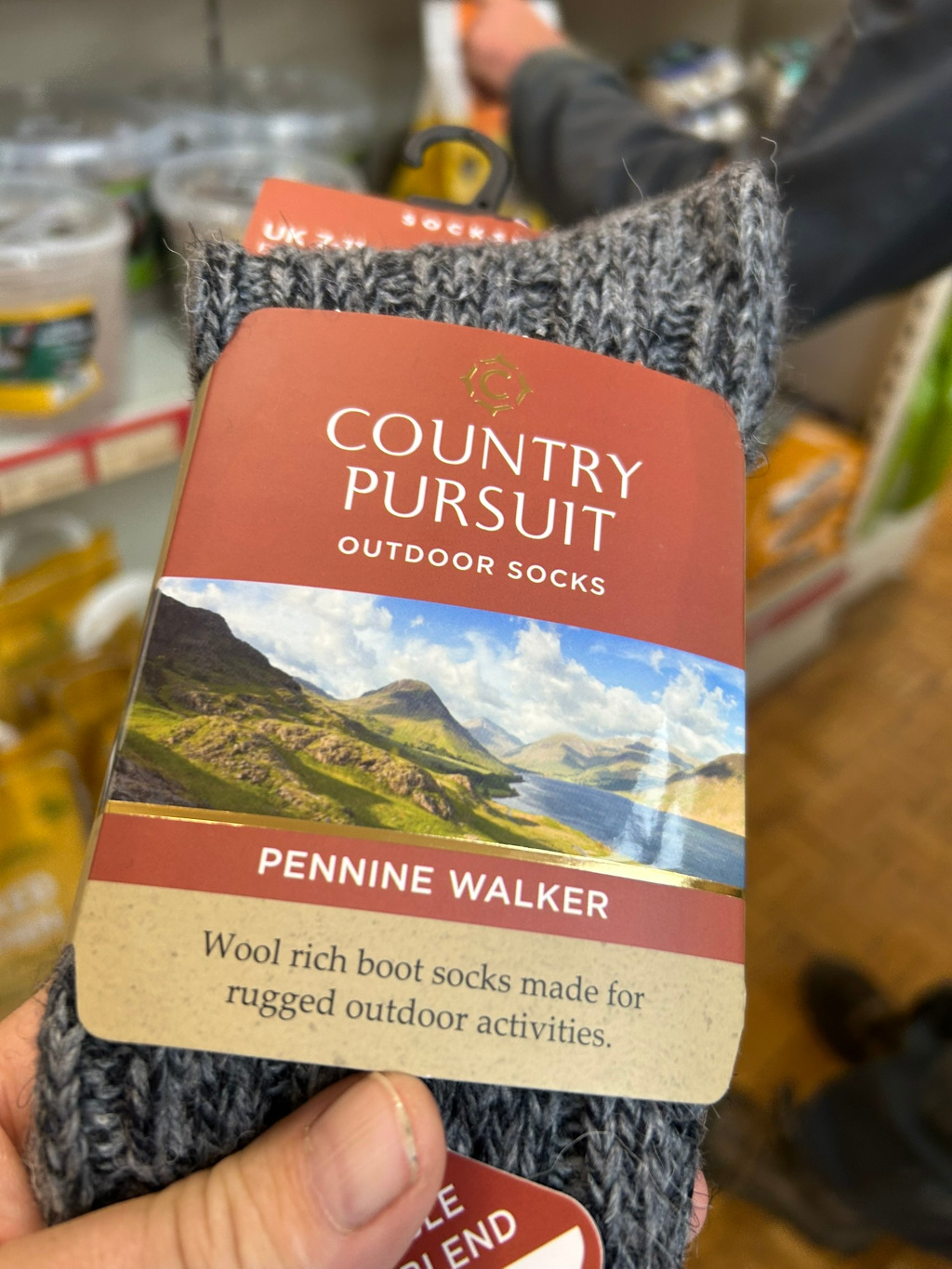 Pair of grey wool hiking socks. The socks are packaged with a tag that advertises them as Country Pursuit Outdoor Socks, specifically the Pennine Walker style. The tag features a scenic image of a mountainous lake district, emphasising the socks suitability for rugged outdoor activities. The overall impression is one of practicality and connection to nature.