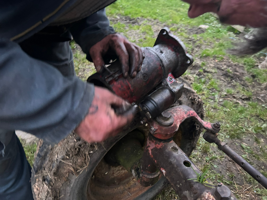 Close-up view of a Charlie's hands working on an old, rusty piece of machinery, possibly a tractor. The individual is wearing dirty work clothes and appears to be meticulously disassembling or repairing a part of the machine's motor or starter. The setting appears to be outdoors in a grassy area.