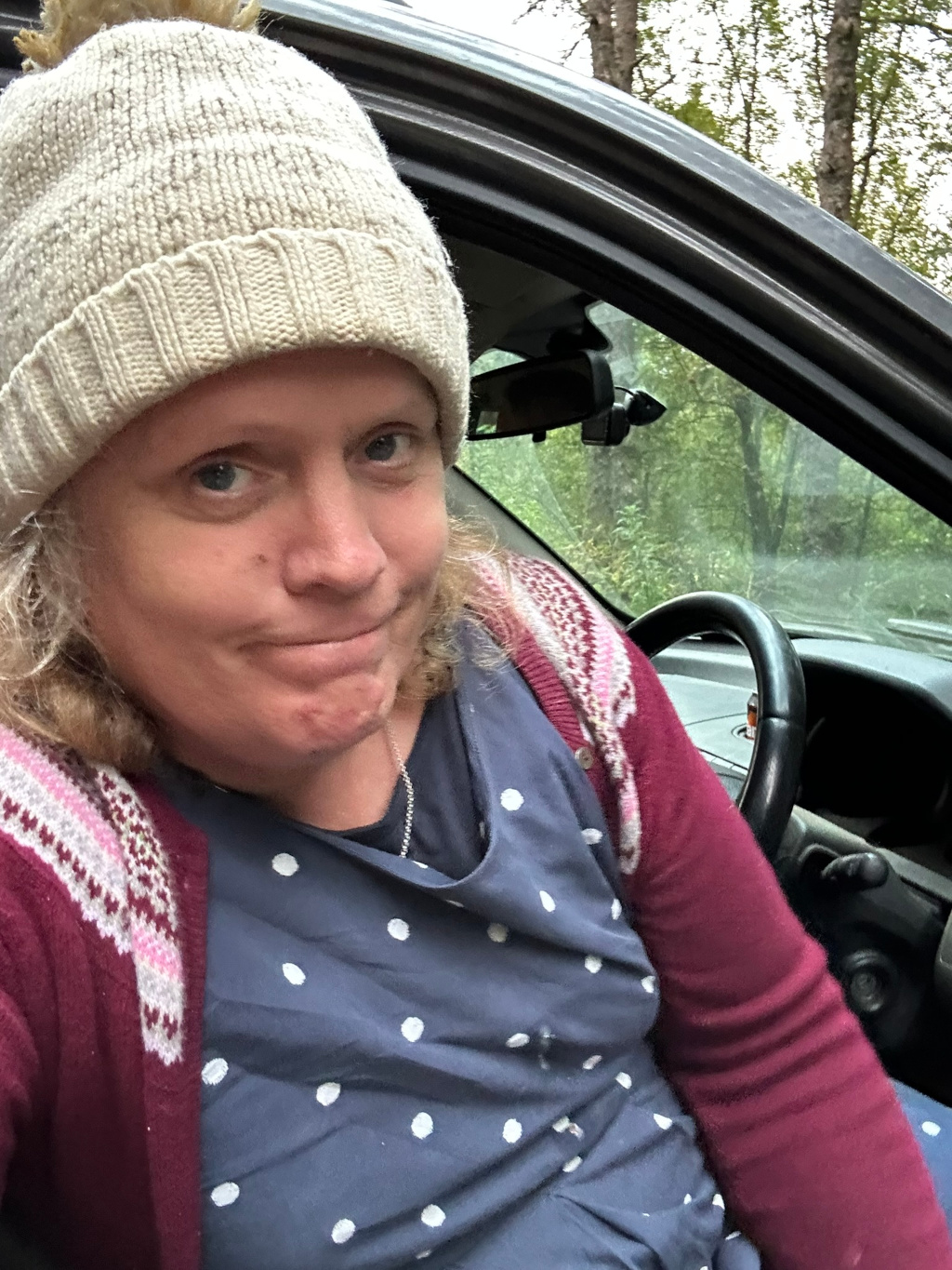 Close-up selfie of a Leonie with fair skin, wearing a cream-coloured knit hat with a small decorative element on top, a dark-blue polka-dotted dress, and a maroon cardigan with a pink and white pattern at the shoulders. Leonie appears to be sitting in the driver's seat of a dark-coloured car, with some trees visible through the window. The expression on Leonie's face is somewhat ambiguous, a slightly quizzical or pensive smirk. The overall impression is one of casual, everyday life.