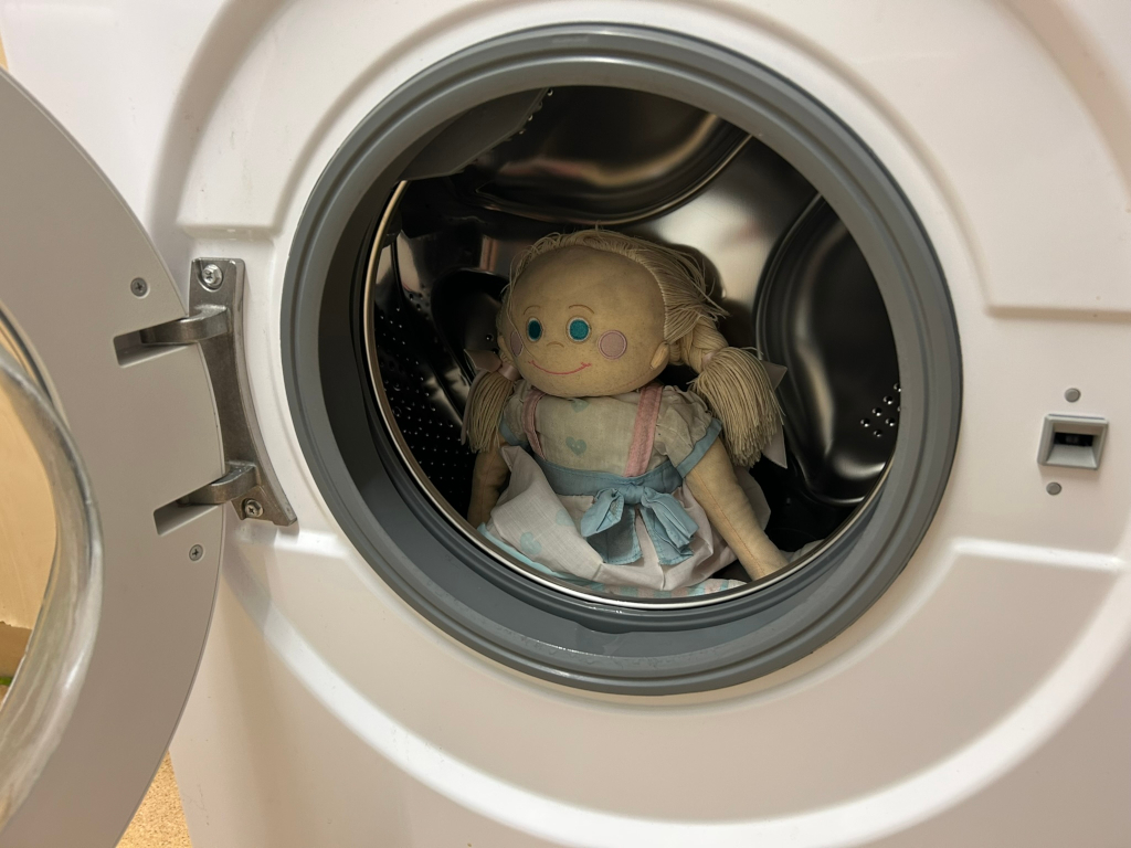 Rag doll sitting inside the drum of a washing machine. The doll is light-skinned with blonde pigtails and a light blue dress. The washing machine is white.
