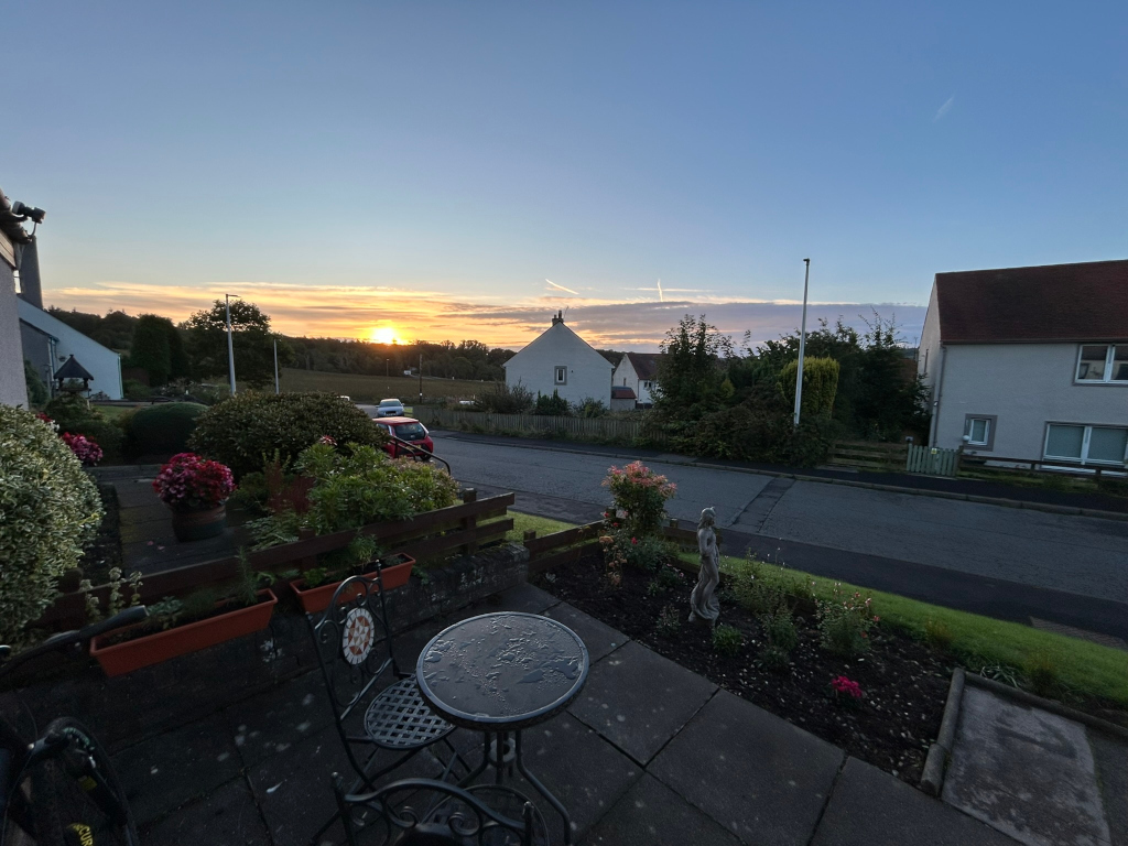 Residential street scene at sunrise. The sun is rising behind a line of trees and houses visible in the mid-ground. In the foreground is a small patio area with a table and chairs, potted plants, and a small garden. The overall mood is peaceful and serene.