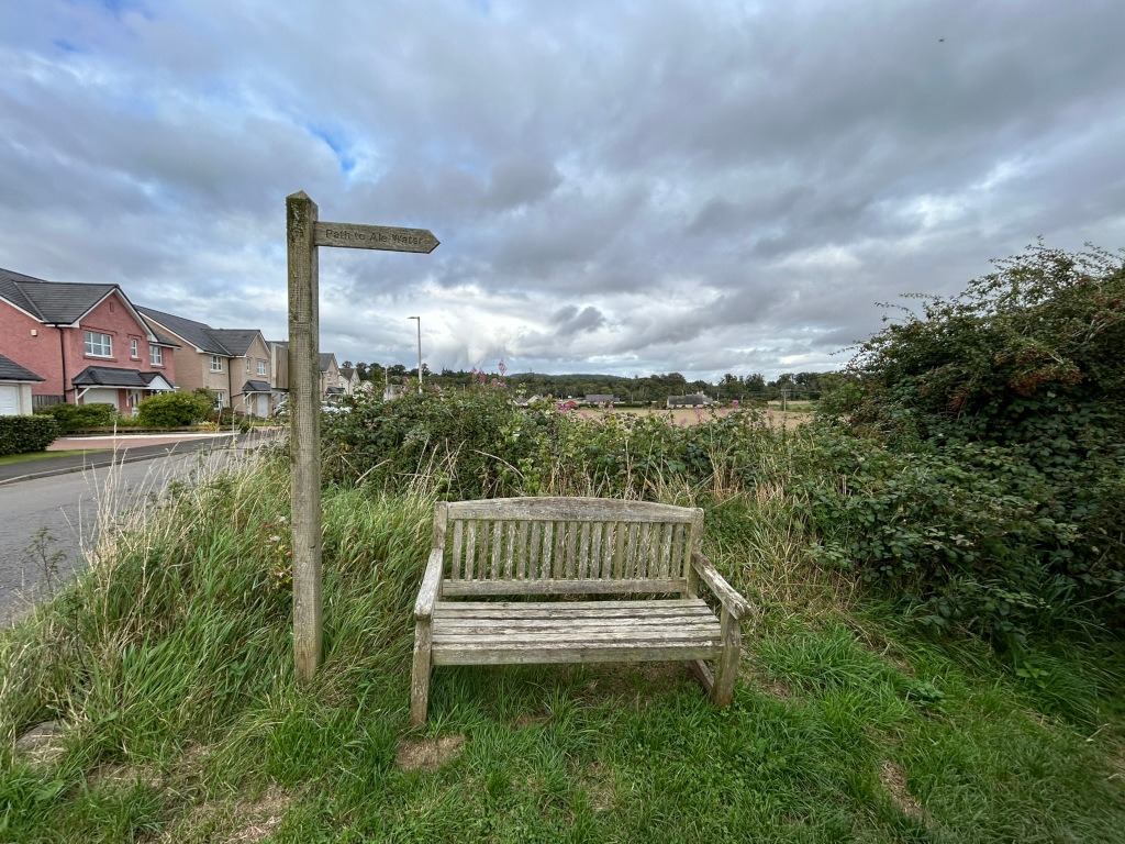 Weathered wooden bench situated beside a wooden signpost that indicates Path to Ale Water. The bench and signpost are positioned at the edge of a grassy verge, with a residential area visible in the background to the left. Beyond the houses, a somewhat flat landscape extends under a cloudy sky. The overall feel is peaceful and somewhat secluded, suggesting a quiet spot for rest along a walking path.