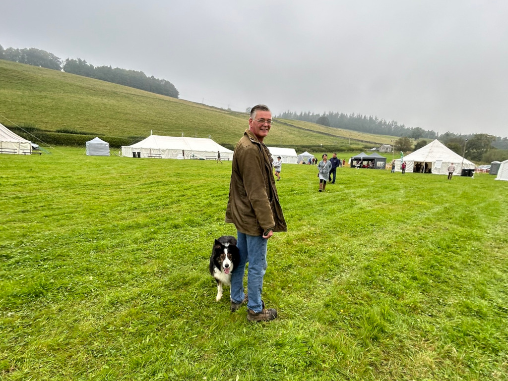 Charlie and his Border Collie dog standing in a grassy field with several white tents in the background. The scene appears to be set at an outdoor event or fair, on an overcast day. Charlie is smiling and looking towards the camera, while his dog is looking towards him. Other people can be seen in the distance, seemingly attending the event. The overall atmosphere is peaceful and relaxed.
