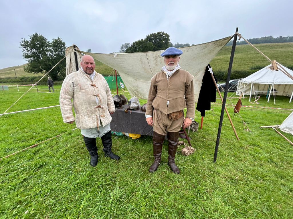 Two men dressed in period clothing, possibly representing 16th or 17th century attire. They stand on a grassy field in front of a makeshift tent structure. One man wears a quilted jacket and tall boots while the other is dressed in a doublet and hose with tall leather boots. Several helmets are visible behind them on a table. The setting suggests a historical re-enactment event or a living history display.