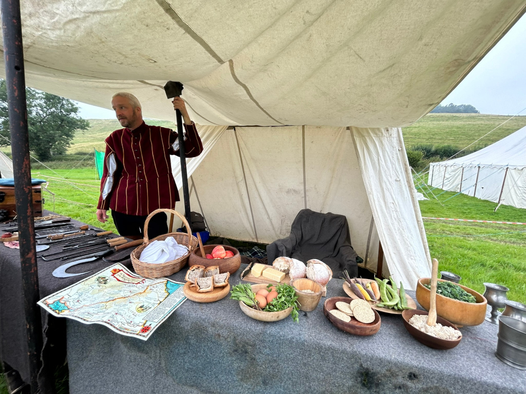 Man in period clothing standing in a canvas tent at what seems to be a historical reenactment event. He is holding a large, dark-colored tool, possibly a type of axe or hammer. A table in front of him is laden with various food items including bread, apples, cheese, vegetables, and what looks like crackers. There are also several metal tools laid out on the table along with a map. The scene is set outdoors in a grassy field. The overall impression is one of a historical demonstration or living history presentation, possibly focusing on a medieval or early modern period.