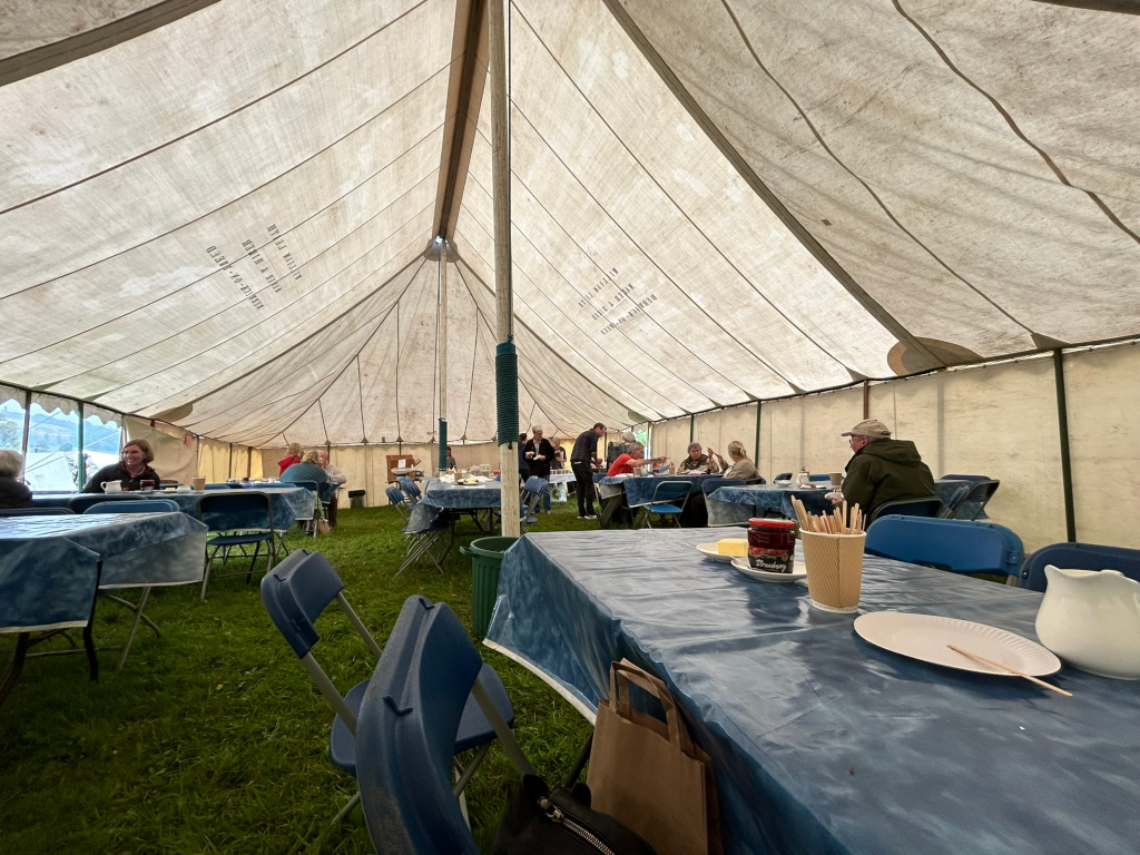Interior of a large canvas tent, likely at an outdoor event. Numerous people are seated at tables covered with blue tablecloths, enjoying a meal or gathering. The atmosphere is casual and communal. The tent's canvas shows signs of age and wear. The setting appears to be a grassy field.