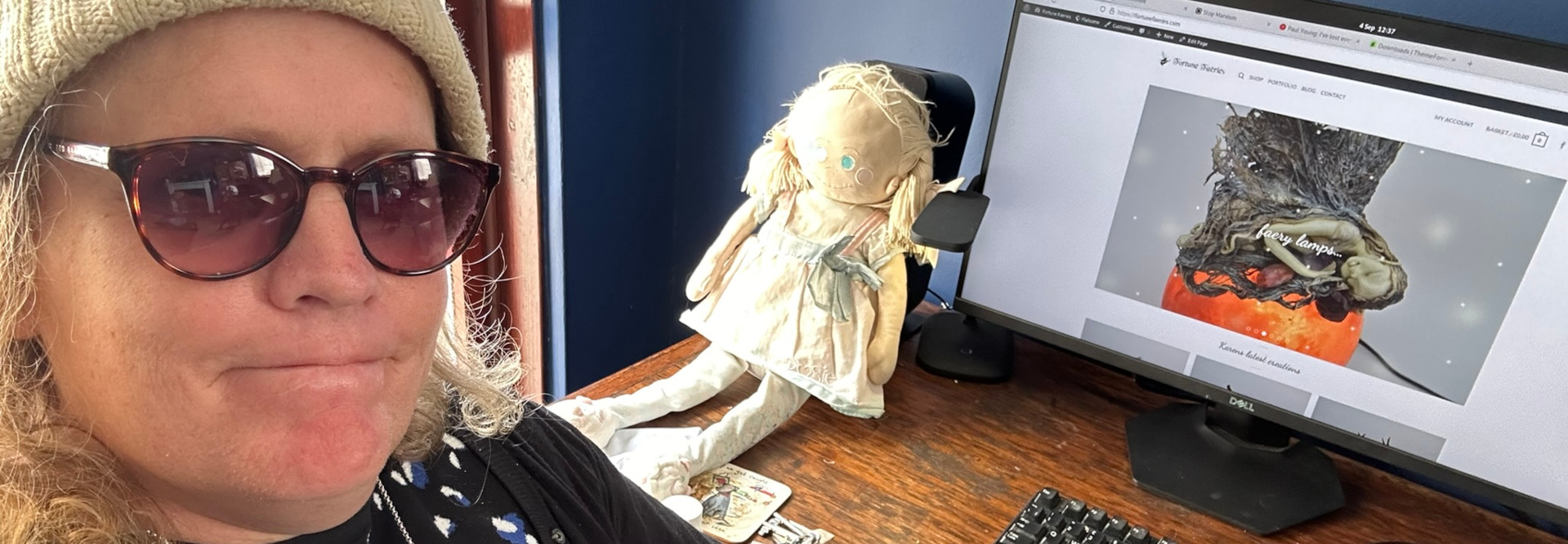 Selfie of a Leonie wearing sunglasses and a knit hat. She is sitting at a desk with a computer displaying a website, which features an image of an artistic lamp seemingly depicting a fairy or nymph figure. A rag doll sits on the desk next to the computer. The overall scene suggests a creative workspace or possibly the artist's personal space, where artistic pursuits and online sales are combined.