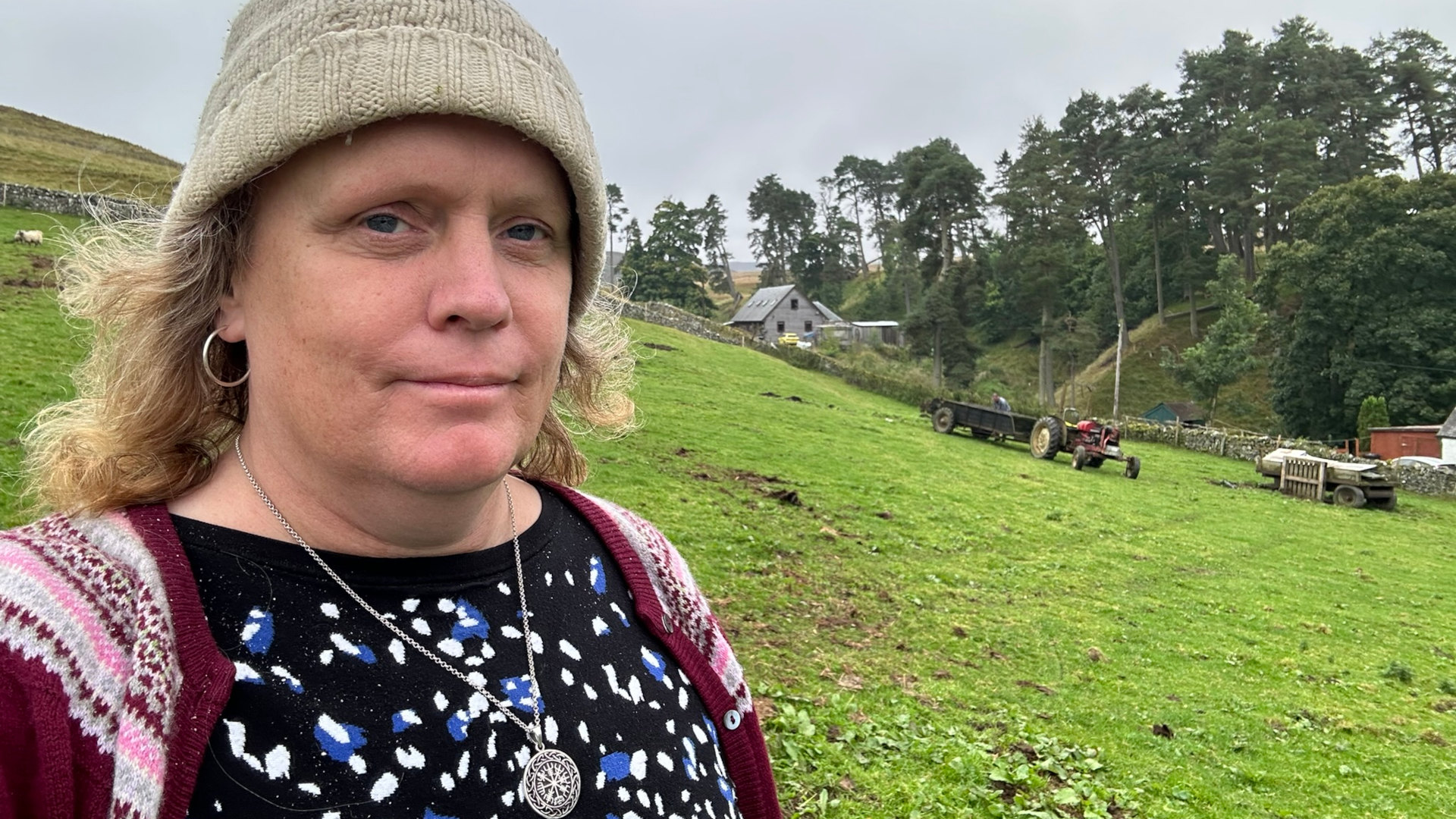 Close-up of a fair-skinned woman with shoulder-length blonde hair, wearing a beige knit hat, a maroon cardigan over a black and blue speckled top, and silver hoop earrings and necklace. She's positioned in the foreground, with a rural landscape visible in the background. This landscape features a grassy field, a stone wall, a farmhouse, and some farm equipment (a red tractor and trailers). A few sheep can be seen in the distance. The overall mood is somewhat contemplative or pensive, given the woman’s expression.