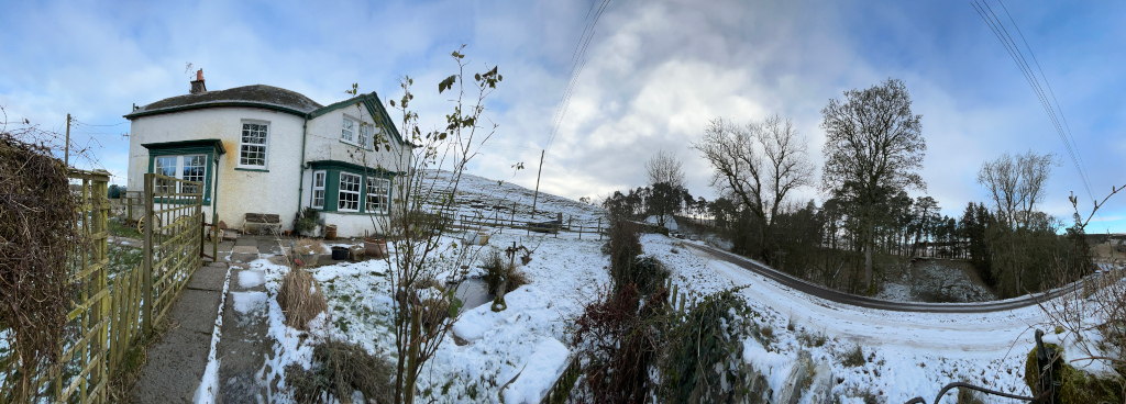 Panoramic view of a snow-covered landscape, centered around a small, white cottage with dark green window frames. The cottage appears quaint and possibly old, nestled amongst a sparsely populated, snow-covered yard. A wooden fence is visible in the foreground, leading toward the cottage. Beyond the cottage, a road curves through a snow-dusted landscape of trees and hills, which seem to be sparsely populated. The sky is a mixture of blue and cloudy, suggesting a cold, winter day. The overall mood is serene and peaceful, evoking a sense of quiet solitude in a rural setting.