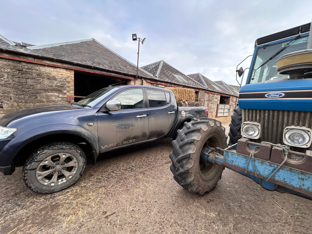 Dark-blue pickup truck, labelled Warrior, parked next to a blue Ford tractor in front of a stone barn. Both vehicles are muddy, suggesting recent agricultural activity. Hay bales are visible inside the barn. The overall scene depicts a rural, working farm setting.