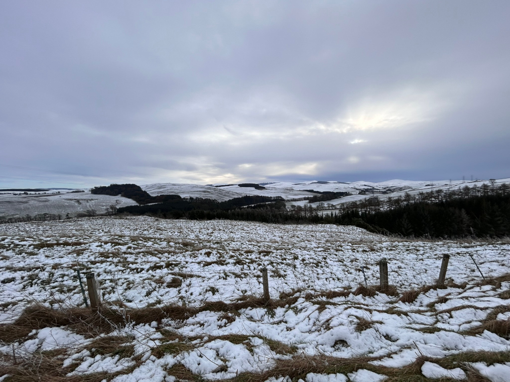 Snow-covered landscape. In the foreground, there is a field of brown grass lightly covered in snow. A wire fence with wooden posts runs across the foreground. In the mid-ground and background, gently rolling hills are visible, also covered in snow. The sky is overcast, a pale grey with some lighter areas suggesting a break in the clouds.