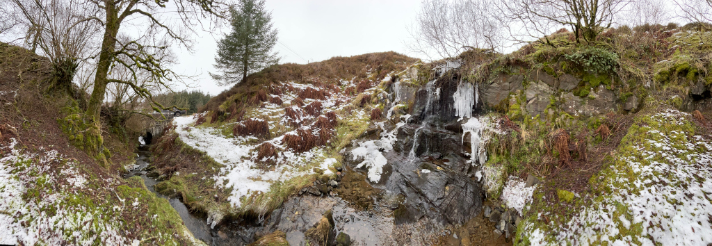 Panoramic view of a small waterfall partially frozen over in a winter landscape. The scene is dominated by rocks, moss, and patches of snow, with a few deciduous trees and a single conifer visible. A small stream leads to the waterfall. The overall impression is one of a serene, cold, and slightly rugged natural setting.
