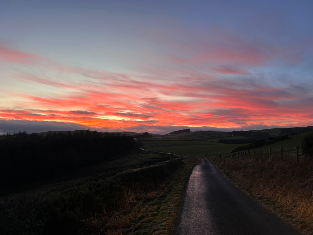 Long, winding road stretching towards a vibrant sunset. The sky is awash in a breathtaking display of pink, orange, and red hues, creating a dramatic contrast against the dark silhouette of rolling hills and a line of trees in the midground. The road appears relatively empty, adding to the sense of tranquillity and vastness. The overall scene evokes a feeling of peace and the beauty of the natural world.