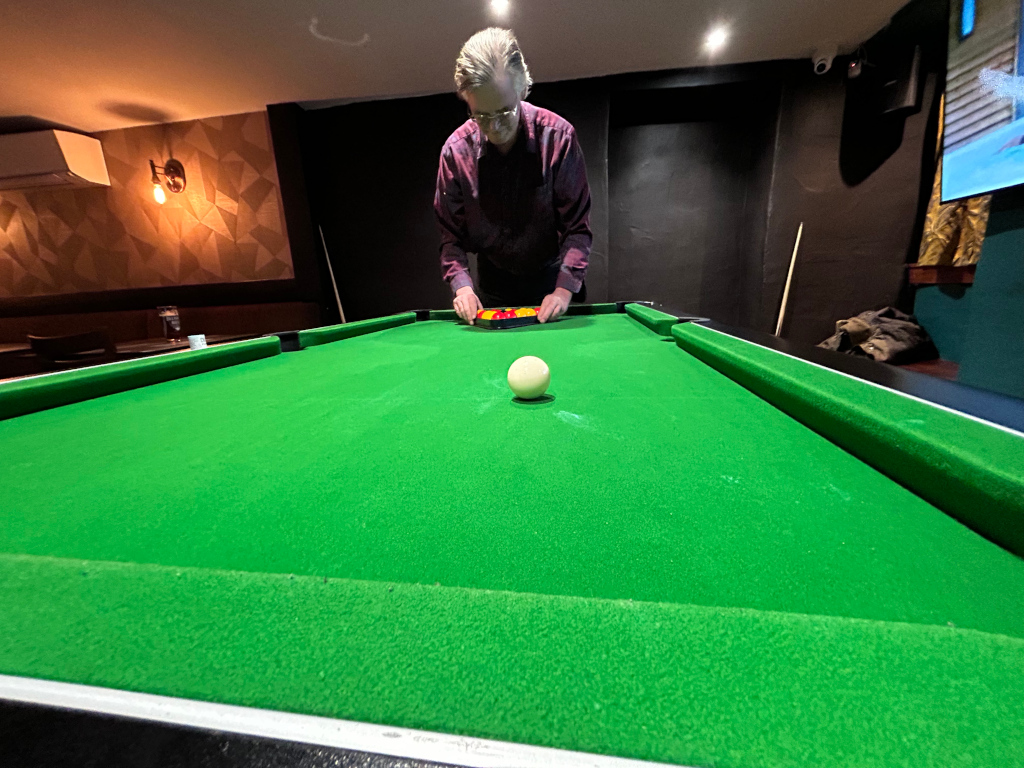 Charlie wearing a burgundy shirt, bending over a pool table. He is holding a pool cue and appears to be about to make a shot. The white cue ball is on the table, near Charlie. The setting appears to be a pub or bar, with a green pool table as the focal point. The background shows some dark wood panelling and geometric wallpaper. A portion of a television screen is visible in the upper right corner.