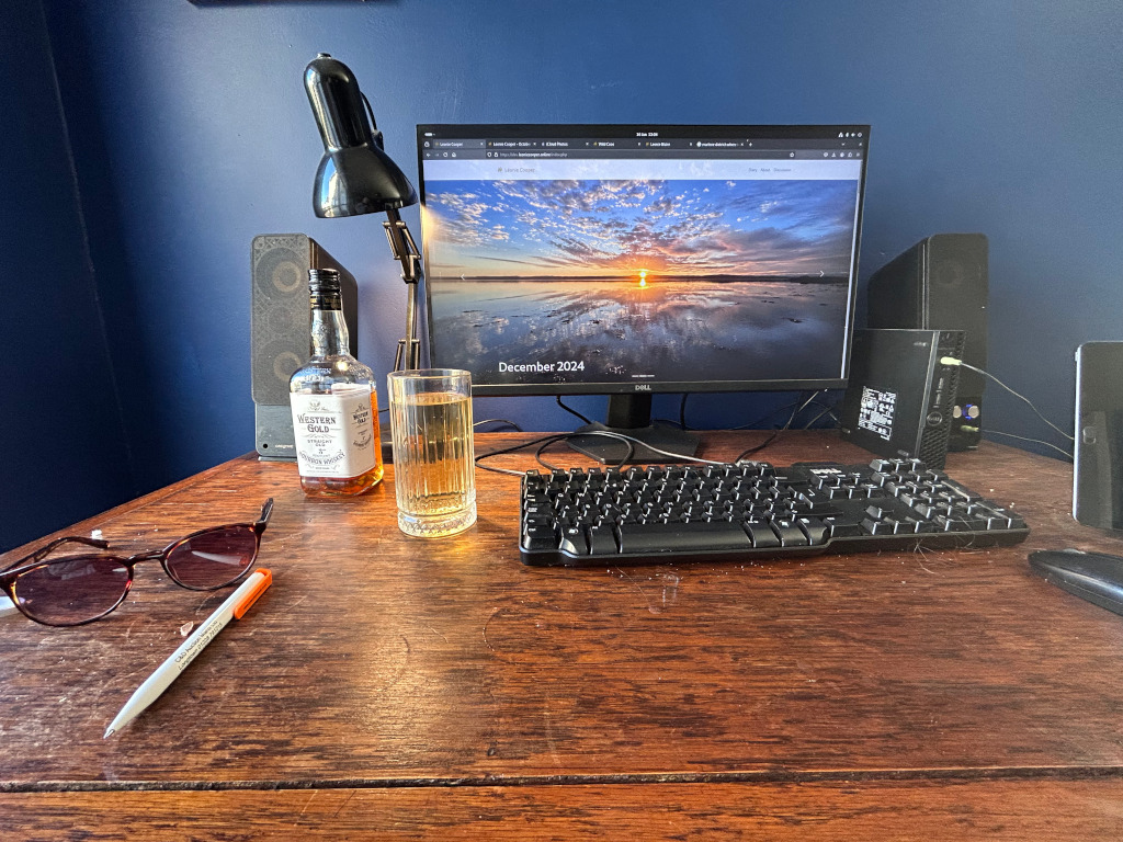 Wooden desk with a computer setup. On the desk is a Dell computer monitor displaying a sunset landscape with the date December 2024, a keyboard, speakers, a bottle of Western Gold Straight Bourbon Whiskey, a glass of what appears to be whisky or similar beverage, a pair of sunglasses, and a pen. The overall ambience suggests a personal workspace or home office.