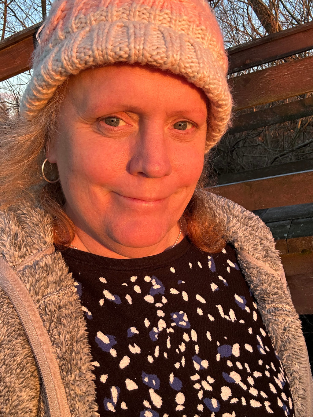 Close-up selfie of a middle-aged woman with blonde hair. She's wearing a chunky knit beanie in shades of off-white and pink, a grey fuzzy jacket, and a dark top with an abstract pattern of off-white and blue shapes. The setting sun casts a warm, orange glow on her face and the wooden structure behind her, possibly a fence or railing. Her expression is somewhat ambiguous, a hint of a smile with a slightly quizzical look in her eyes.