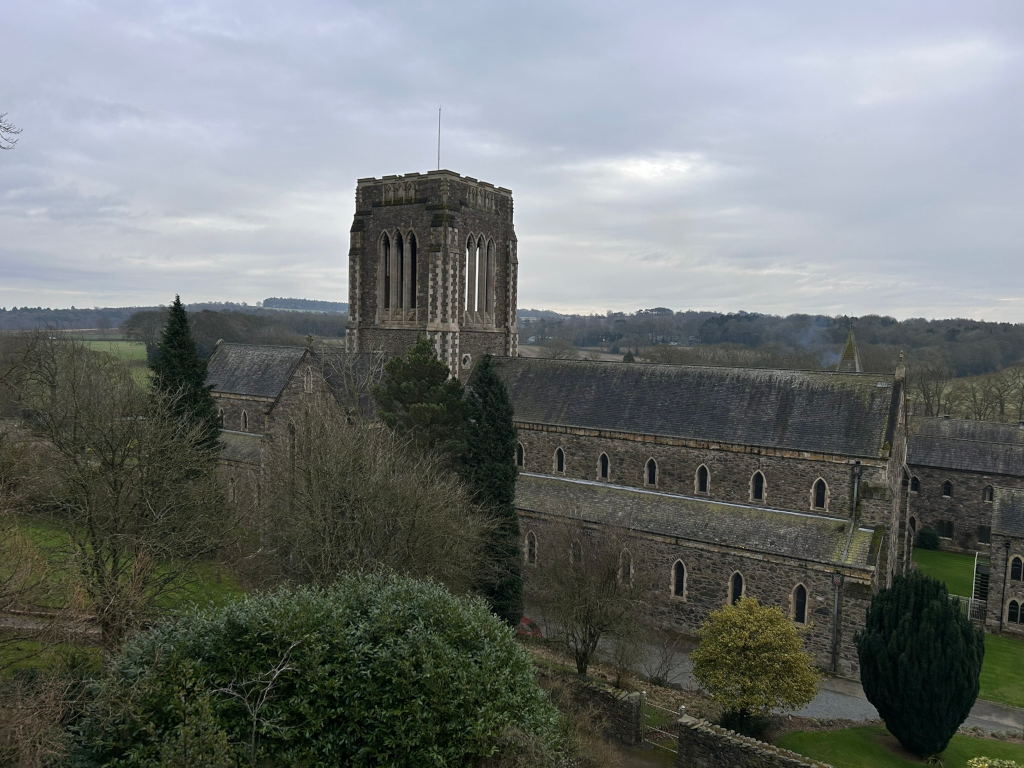 Historical stone abbey set against a backdrop of a gently rolling, tree-covered landscape under a cloudy sky. The abbey itself is the main focus, showcasing its grey stone architecture, long, rectangular structure, and a prominent square tower. The architectural style suggests a medieval or perhaps early modern period. The roof is dark grey slate, and numerous arched windows are visible along the side of the building. The surrounding landscape is peaceful and relatively muted in colour, with a mix of deciduous trees appearing bare or winter-dormant in the foreground and a more distant, darker line of trees on the horizon. The sky is overcast, providing a soft, diffuse light. The overall mood is one of quiet contemplation and historical weight.