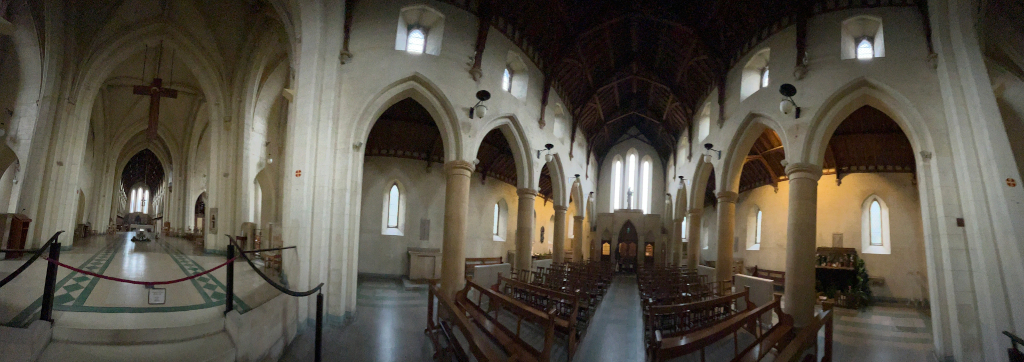 Panoramic view of the interior of an abbey. The architecture is Gothic Revival style, characterised by pointed arches, tall columns, and a long nave leading to an altar. Wooden pews are arranged facing the altar. A large wooden crucifix hangs near the ceiling. The overall atmosphere is serene and reverent, typical of a place of worship.