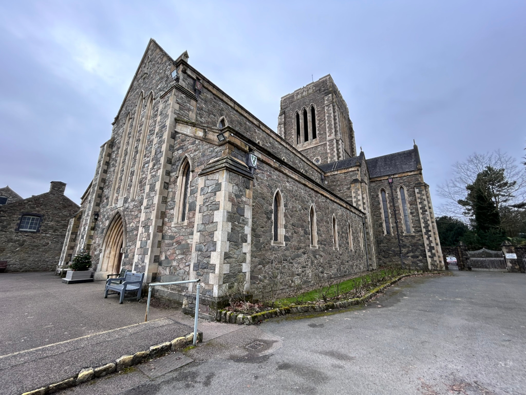Side view of a large stone abbey. The abbey is built of grey stone, with a tall square tower at one end. The style appears to be Gothic Revival. A smaller, adjacent building is partially visible in the background to the left. There's a simple paved area in front of the abbey, with a couple of benches and some landscaping. The sky is overcast. The overall impression is one of quiet dignity and historical significance.