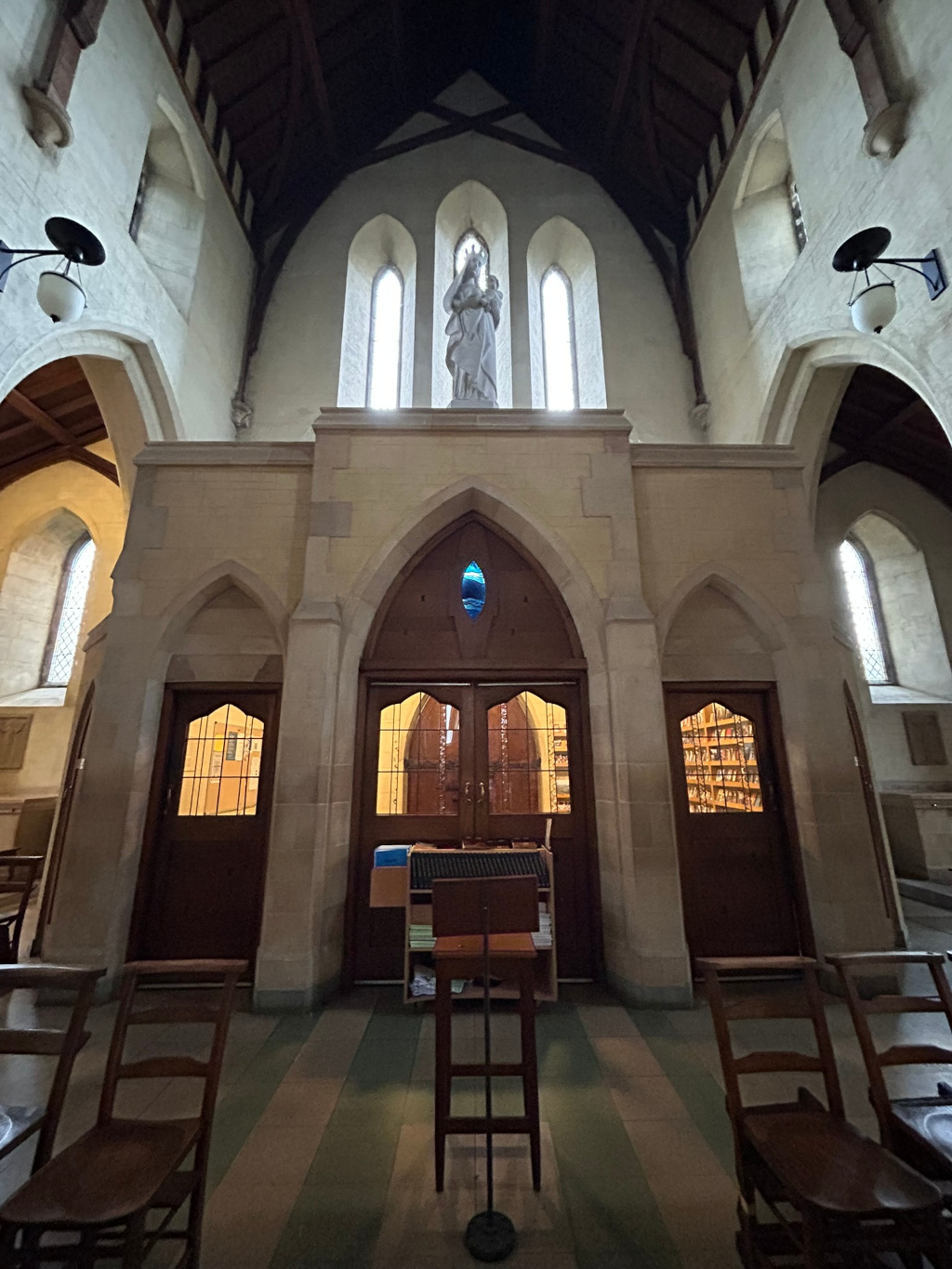 Interior of an abbeys sanctuary. A statue of the Virgin Mary and Child is prominently displayed on a raised altar at the back. Below the statue, a large arched alcove contains a pair of wooden doors leading to another area within the church presumably a sacristy or chapel. Flanking this alcove are smaller, similarly arched recesses, each containing a wooden door, possibly leading to storage or smaller chapels. The floor is tiled, and wooden chairs are visible in the foreground. The overall architectural style is Gothic Revival, characterised by pointed arches and stained glass windows. The lighting focuses on the central statue, creating a contemplative and solemn atmosphere.