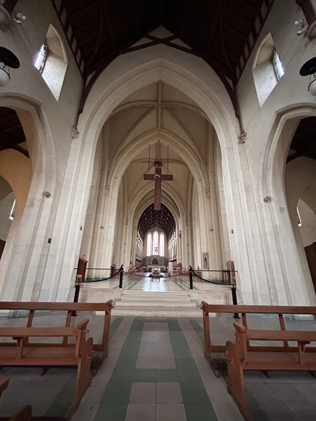 nave of an abbey, looking down its length towards the altar. The architecture is Gothic Revival, featuring high, pointed arches, tall columns, and a large, wooden crucifix hanging from the ceiling. The floor is tiled, and simple wooden benches are placed near the viewer. The overall feeling is one of quiet solemnity and grandeur.