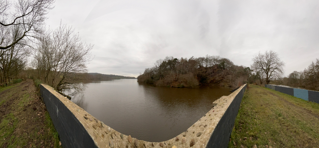 Panoramic view of a calm reservoir. The water is dark brown and relatively still. The lake is bordered by bare, winter trees on both sides, creating a quiet, somewhat desolate atmosphere. A low stone wall, partially covered with dark material, is visible in the foreground, suggesting a dam or embankment. The sky is overcast and grey. The overall impression is one of a peaceful, natural scene in the winter season.