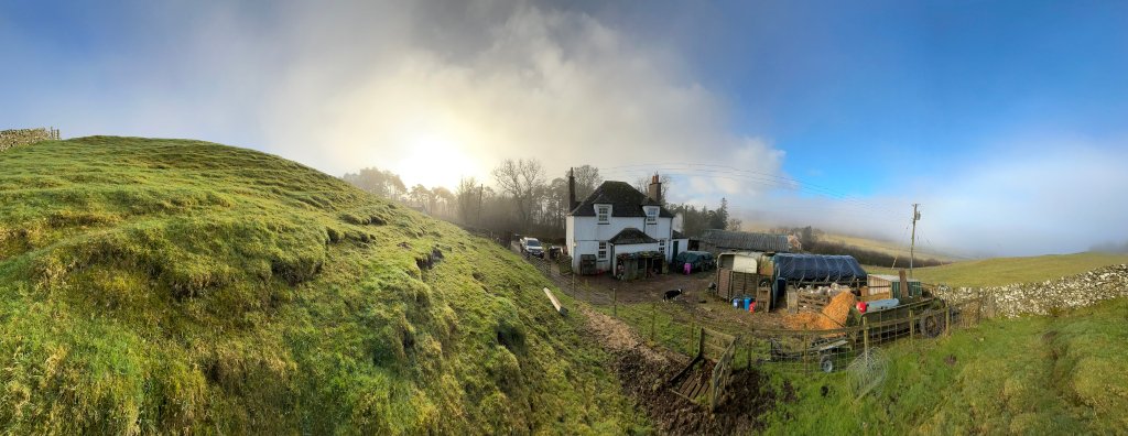 Panoramic view of a rural scene. In the foreground is a grassy hillside, leading to a white farmhouse situated in a farmyard. Various farm implements and structures, including what appears to be a trailer and a large tarp, are visible in the yard. A low stone wall is partially visible in the background, and a misty or foggy landscape extends into the distance under a partly cloudy sky. The overall impression is one of a serene and somewhat isolated farm setting.