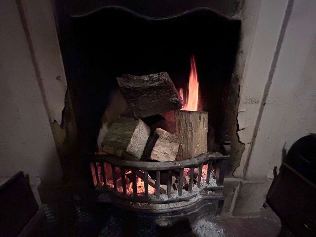 Fire burning in a traditional fireplace. The fire is relatively small, with several pieces of wood burning within a metal grate. The fireplace itself appears old, with some visible damage or wear to the surrounding wall. The overall atmosphere is dark and somewhat dimly lit, focusing attention on the flickering flames.