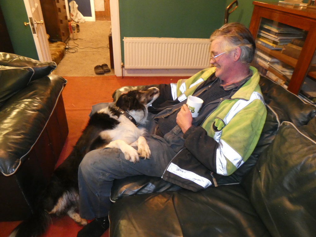 Charlie relaxing on a leather couch with his border collie dog. Charlie is wearing a lime green high-visibility jacket and is holding a mug. The dog is nestled comfortably on his lap. The setting appears to be a living room. The overall feeling is one of quiet companionship and relaxation.