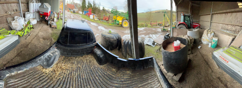 Panoramic view of a farmyard scene, possibly taken from inside a barn. A dark-coloured pickup truck with a canopy is parked in the foreground, inside a structure that appears to be a barn or shed. The truck bed contains some hay. Surrounding the truck are various farm implements, including large bags of what looks like feed or fertiliser, large tires, and other pieces of agricultural equipment. There are also several containers, some with grain or feed stuff. In the background, outside the structure, more farm machinery is visible. The setting appears to be rural, with fields and trees in the distance. The overall impression is one of a busy, working farm environment, with equipment and supplies stored both inside and outside a barn.