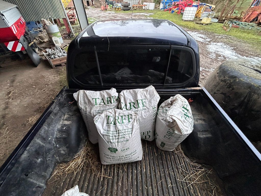 Back of a dark-coloured pickup truck parked in a farmyard. The truck bed contains several large bags of Tarff feed, a type of animal feed as indicated by the branding on the bags. The farmyard setting is evident from the surrounding agricultural equipment and structures visible in the background. The overall impression is one of routine farm work or chores. There is no apparent narrative or deeper moral message; it is a straightforward depiction of a common agricultural task.