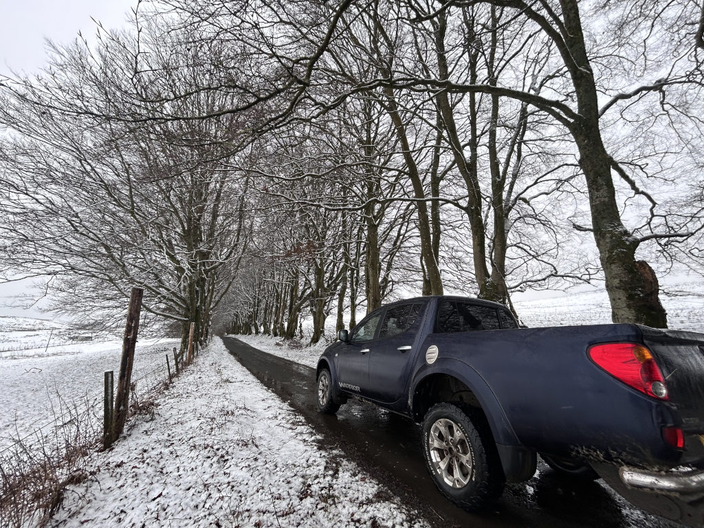 Dark blue pickup truck parked on a snowy, narrow road lined with leafless trees covered in a light dusting of snow. The scene is quiet and wintry, with a muted colour palette dominated by whites, grays, and the dark blue of the vehicle. The road appears to lead into the distance, disappearing between the trees. The overall impression is one of peaceful solitude in a snowy landscape.