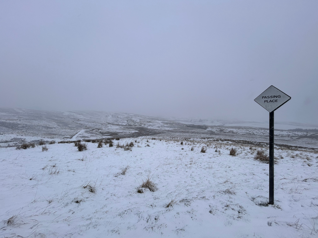 Snow-covered landscape with a single Passing Place sign. The sky is overcast and the overall atmosphere is bleak and cold. The sign stands alone in the snowy expanse, suggesting a sense of isolation or perhaps the anticipation of an encounter.