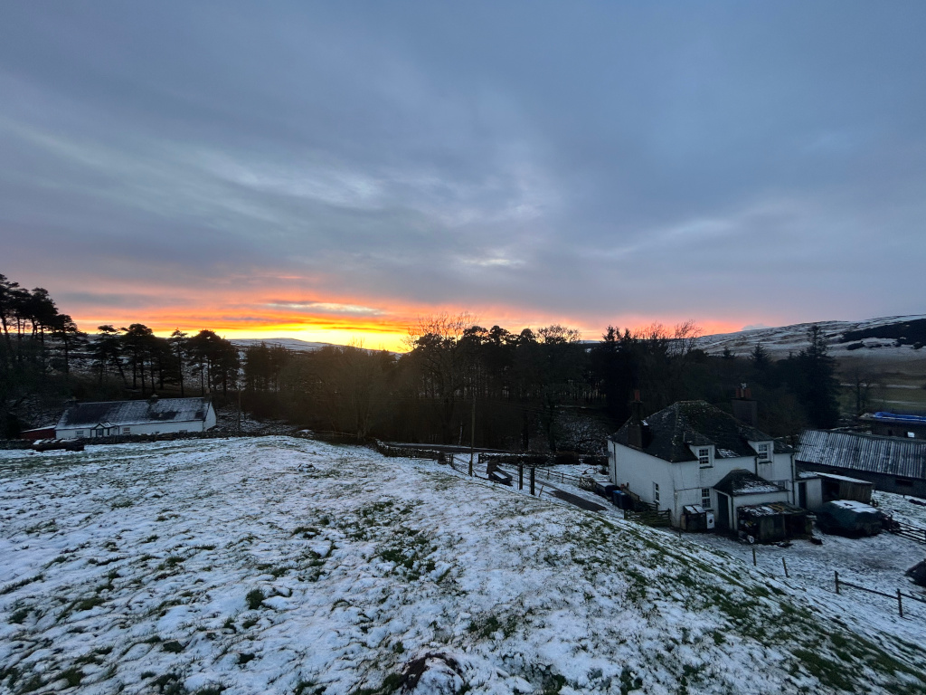 Snow-dusted landscape at sunset. In the foreground, a gently sloping hill covered in a thin layer of snow leads to two houses. The closest house is a white two-story building, while a larger, lower building is slightly further away. Behind the houses, a line of dark evergreen trees stretches across the middle ground. The background reveals rolling hills under a dramatic sunset sky, with warm orange and pink hues blending into cooler blues and grays. The overall mood is serene and peaceful, capturing the beauty of a rural winter sunset.