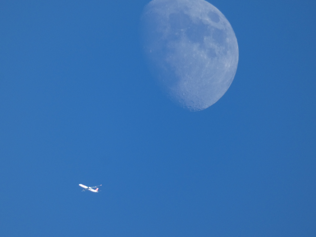 Partially visible moon in the upper right quadrant of a clear blue sky. A small airplane is visible in the lower left quadrant, flying horizontally. The vastness of the sky and the relative sizes of the moon and aeroplane create a sense of scale and distance.