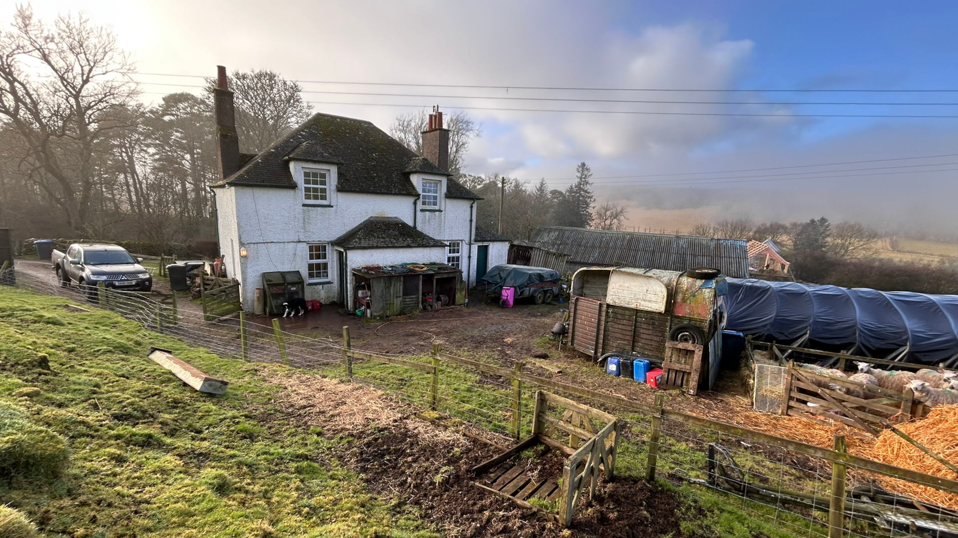 Rural scene featuring a whitewashed farmhouse at the centre. The farmhouse is surrounded by farm animals (sheep are visible in a pen), farm equipment (including a horse trailer and a pickup truck), and various outbuildings. The overall impression is one of a working farm, possibly a sheep farm, on a slightly misty morning. The scene exudes a quiet, peaceful, and somewhat rustic atmosphere. The background shows rolling hills and farmland, extending into a light fog or mist.